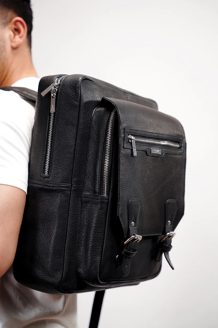 img_6a0c58c0db5e87.51464975 Black leather backpack with buckle and zipper closures, worn by a person